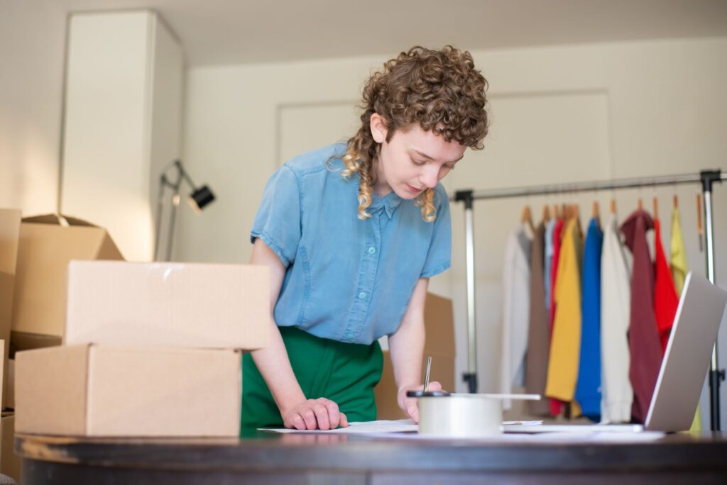 Emprendedora de una tienda en línea revisando pedidos junto a cajas de envío, una laptop y un perchero con ropa
