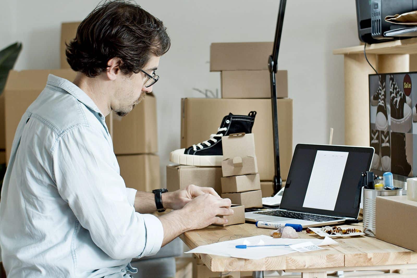 Emprendedor de una tienda en línea preparando paquetes junto a una laptop con una hoja de pedidos, escena típica de pedidos que se pasan a mano